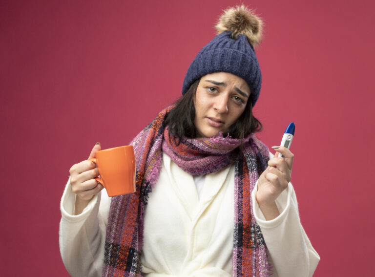 weak-young-caucasian-ill-girl-wearing-robe-winter-hat-scarf-holding-cup-tea-thermometer-looking-camera-isolated-crimson-background