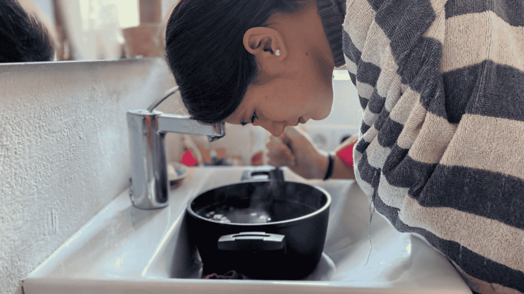 Mujer inclinada sobre una olla con agua caliente inhalando vapor, cubierta con un poncho a rayas para aliviar problemas respiratorios.
