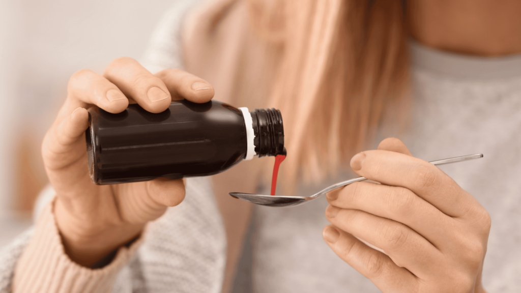 Persona sirviendo un jarabe rojo desde un frasco oscuro hacia una cuchara, lista para su consumo, con fondo desenfocado.